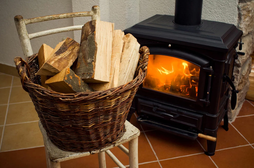 A basket with wood logs next to a stove. 