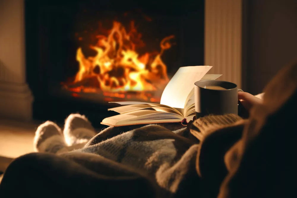Woman with a cup of drink and book near fireplace. 